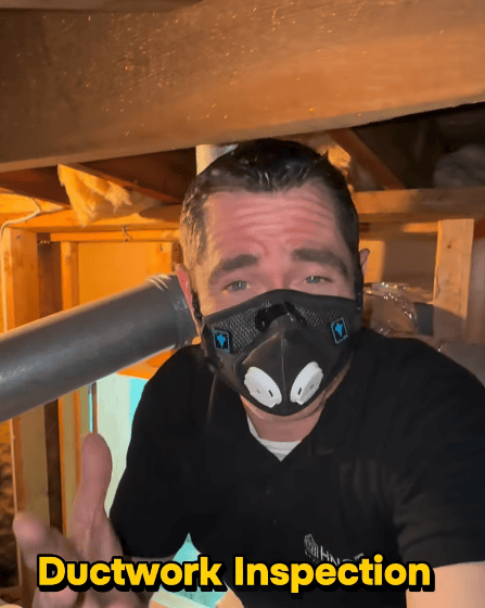 Inspector wearing protective equipment inside a crawlspace beneath a home, near flexible HVAC ductwork.