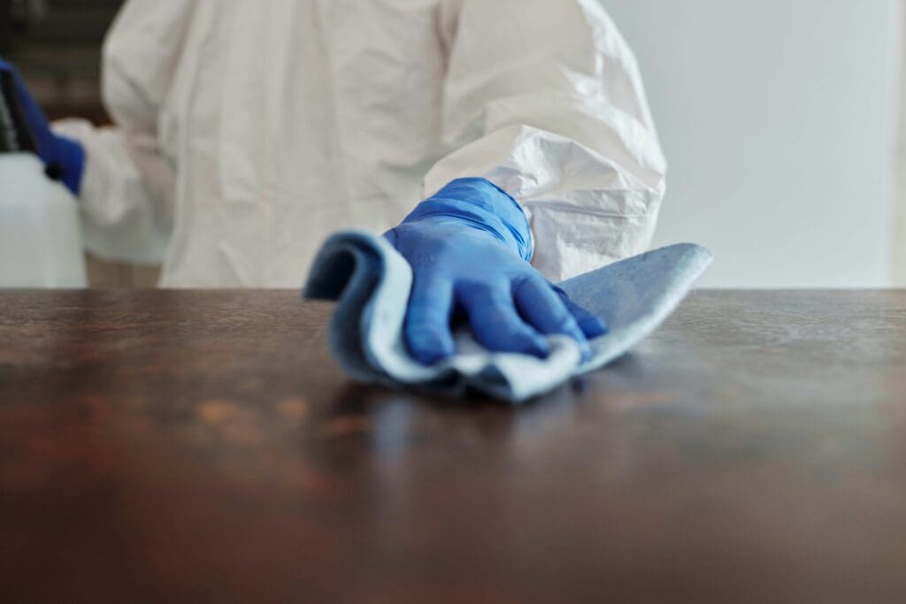 A person wearing protective clothing and blue gloves wipes a wooden table with a white cleaning cloth.